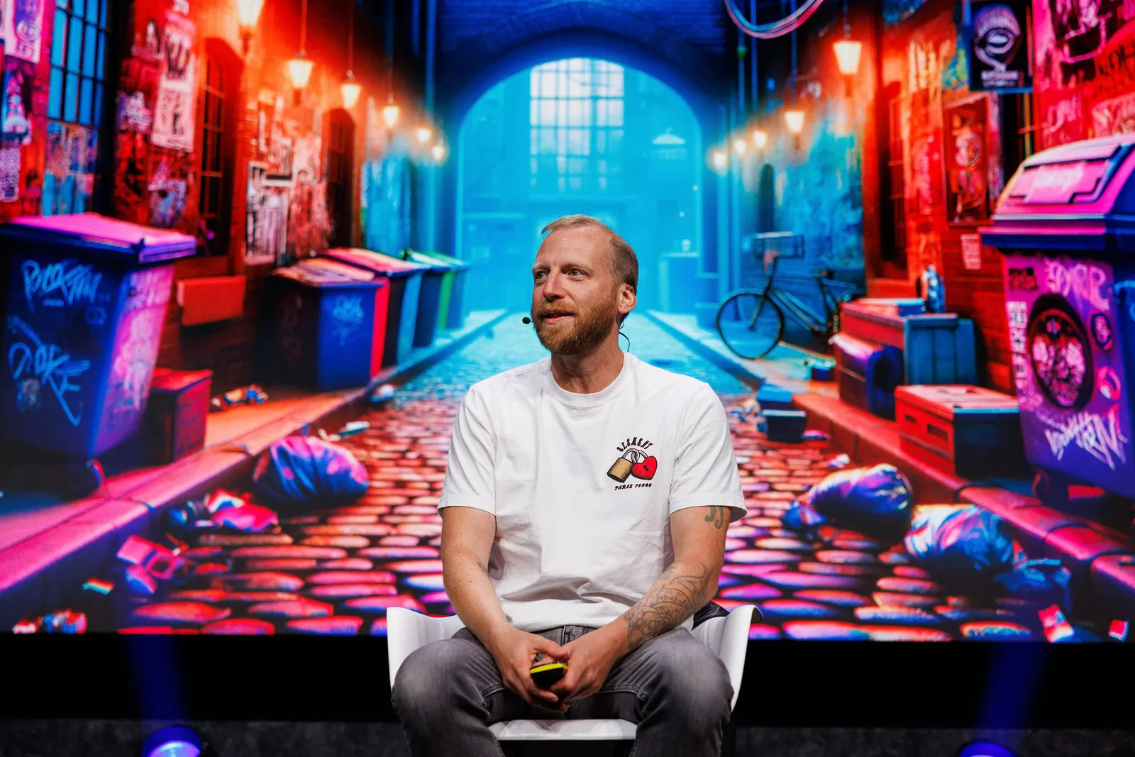Parry Malm on stage at AntiCon, wearing a headset microphone in front of a large screen showing a neon-lit graffiti alleyway.
