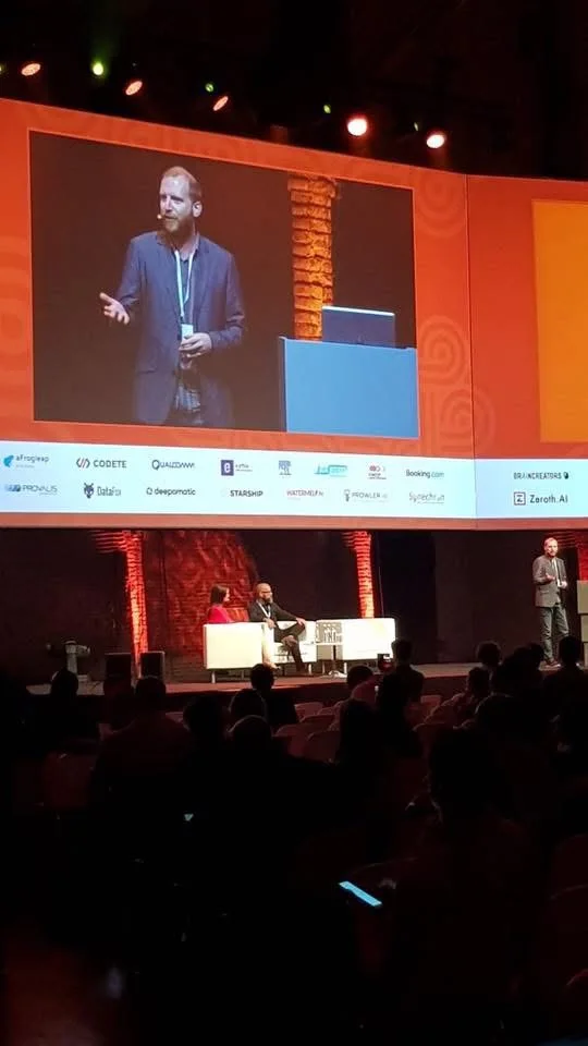 Parry Malm speaking on a main stage, his face projected on a large screen above him, in front of a full auditorium.